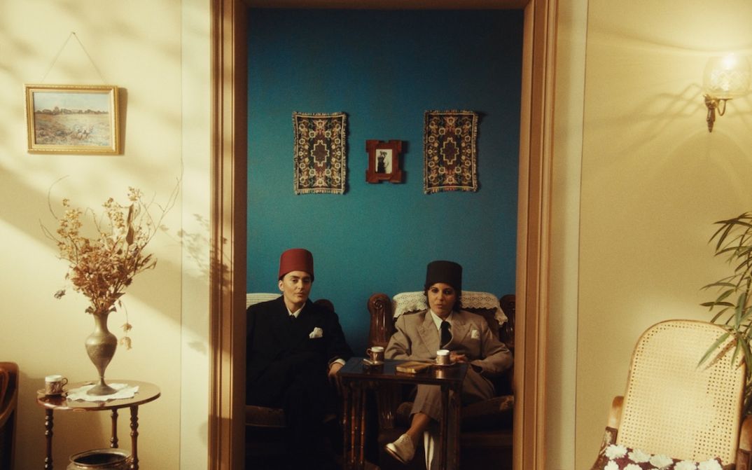 Filmstill aus NEO NAHDA: Zwei als Männer gekleidete Frauen sitzen zwischen einem Türrahmen und blicken direkt in die Kamera.