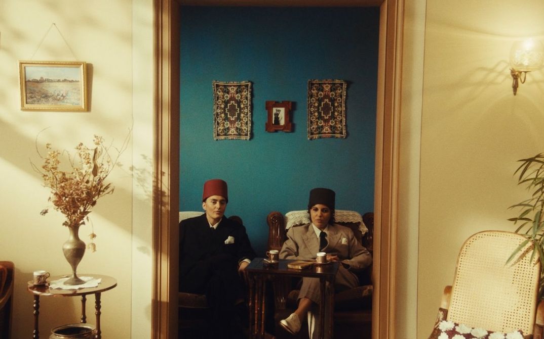 Filmstill aus NEO NAHDA: Zwei als Männer gekleidete Frauen sitzen zwischen einem Türrahmen und blicken direkt in die Kamera.