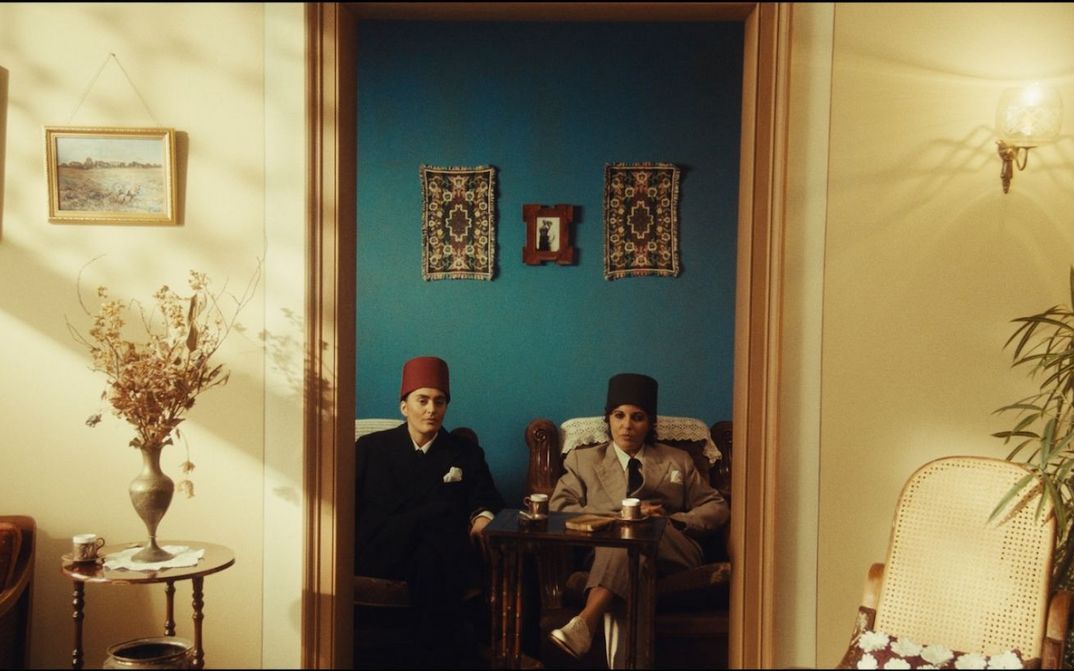Film still from NEO NAHDA: Two women dressed as men sit between a door frame and look directly into the camera.