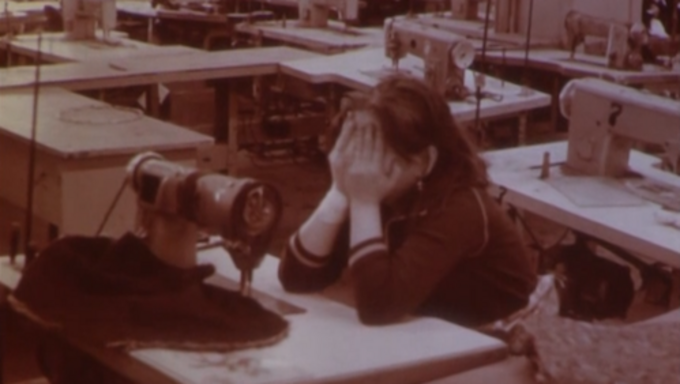 Film still from WOMEN BEHIND A STONE WALL: In a room full of sewing tables, a woman sits with her face buried in her hands.