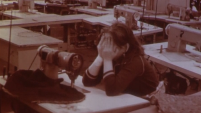 Filmstill aus WOMEN BEHIND A STONE WALL: In einem Raum voller Nähtische sitzt eine Frau und vergräbt ihr Gesicht in den Händen.