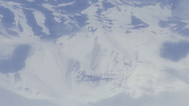 Filmstill aus dem Film „detours while speaking of monsters“ von Deniz Şimşek. Ein Blick aus der Vogelperspektive auf eine entsättigte, schneebedeckte Berglandschaft. 