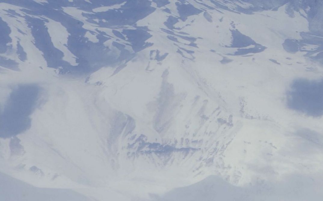 Filmstill aus dem Film „detours while speaking of monsters“ von Deniz Şimşek. Ein Blick aus der Vogelperspektive auf eine entsättigte, schneebedeckte Berglandschaft.