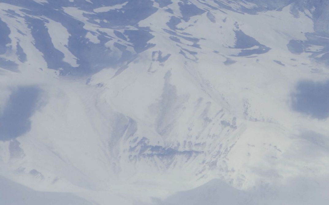 Film still from Deniz Şimşek’s film “detours while speaking of monsters”. A birds-eye view of a desaturated, snow-covered mountain landscape.