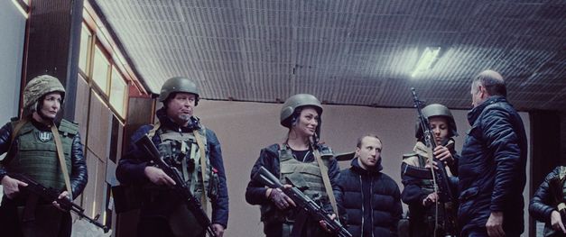 It shows an indoor area with a ceiling lamp and covered windows. 5 people in military protective clothing stand next to each other and receive rifles from a man.