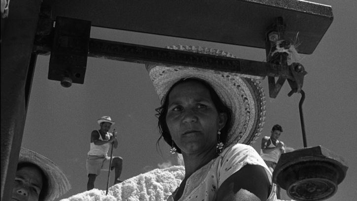 Film still from ARAYA: A woman with a straw hat in the foreground. In the background are three other people standing on a hill.