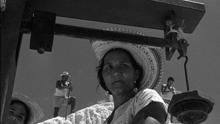 Film still from ARAYA: A woman with a straw hat in the foreground. In the background are three other people standing on a hill.