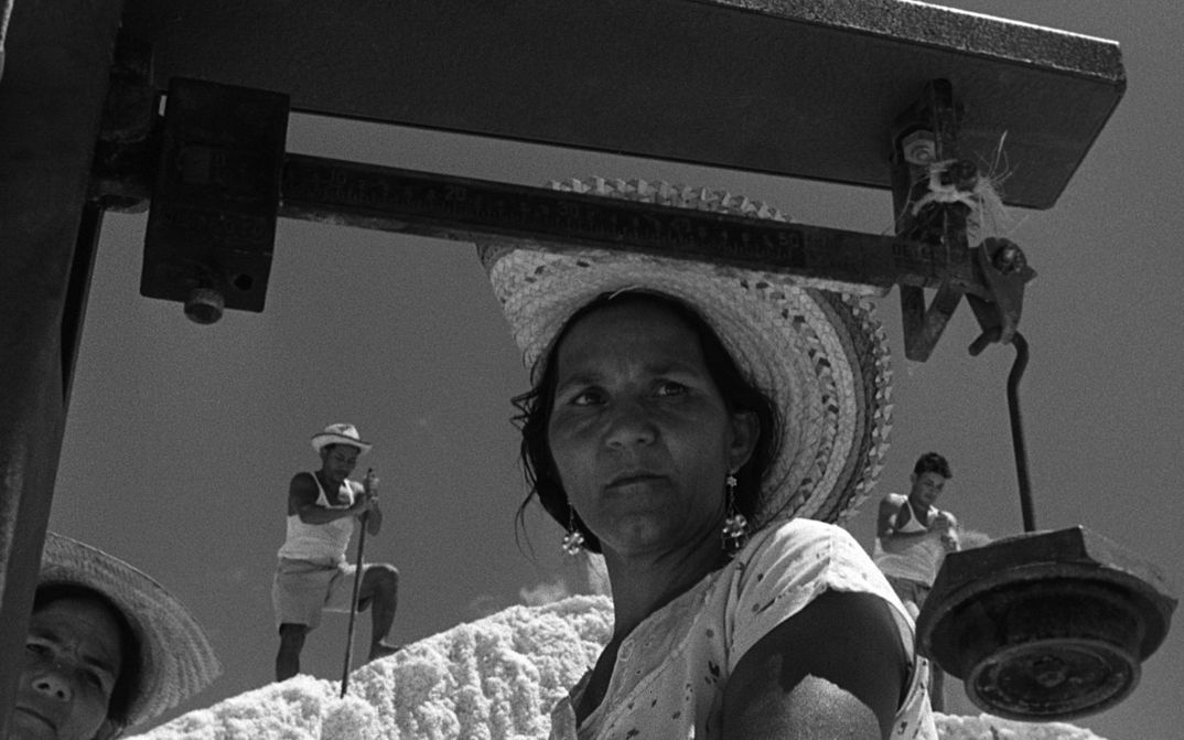 Film still from ARAYA: A woman with a straw hat in the foreground. In the background are three other people standing on a hill.