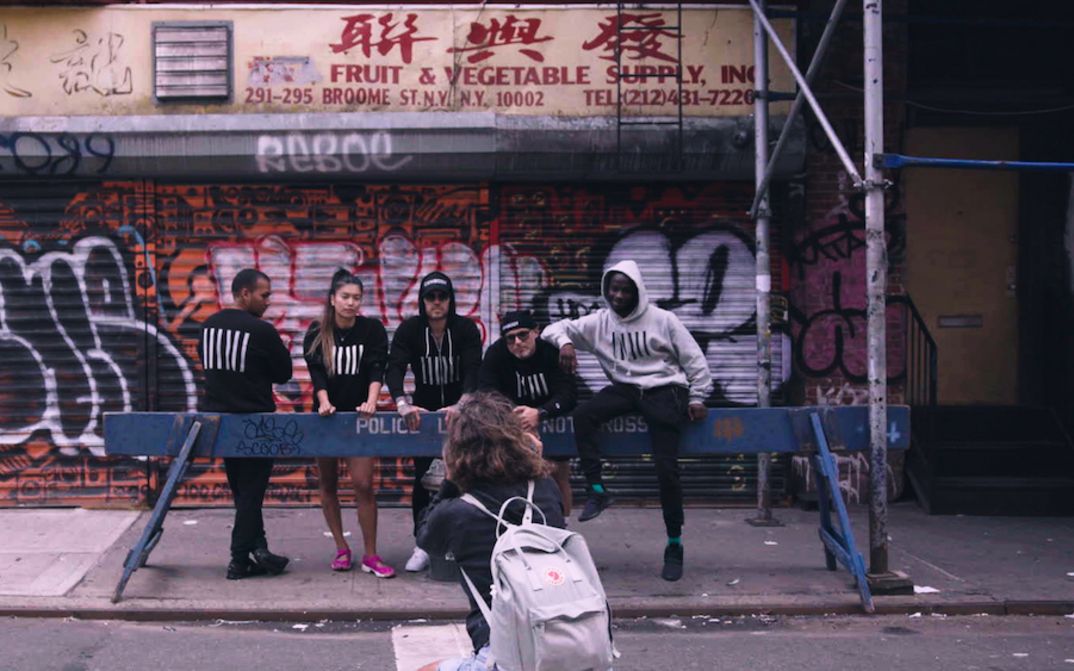 Film still from CONBODY VS. EVERYBODY: Five people, wearing matching sweatshirts, are posing for a photographer on a New York street corner in front of a police barrier.