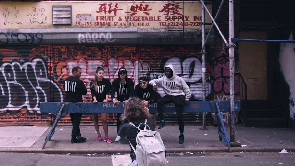 Film still from CONBODY VS. EVERYBODY: Five people, wearing matching sweatshirts, are posing for a photographer on a New York street corner in front of a police barrier.
