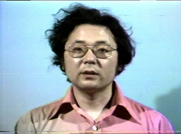 Still from “I Am (Not) Takahiko Iimura, I Am (Not) Akiko Iimura”: a man in a pink shirt, glasses and black hair (the artist himself) looks directly into the camera.