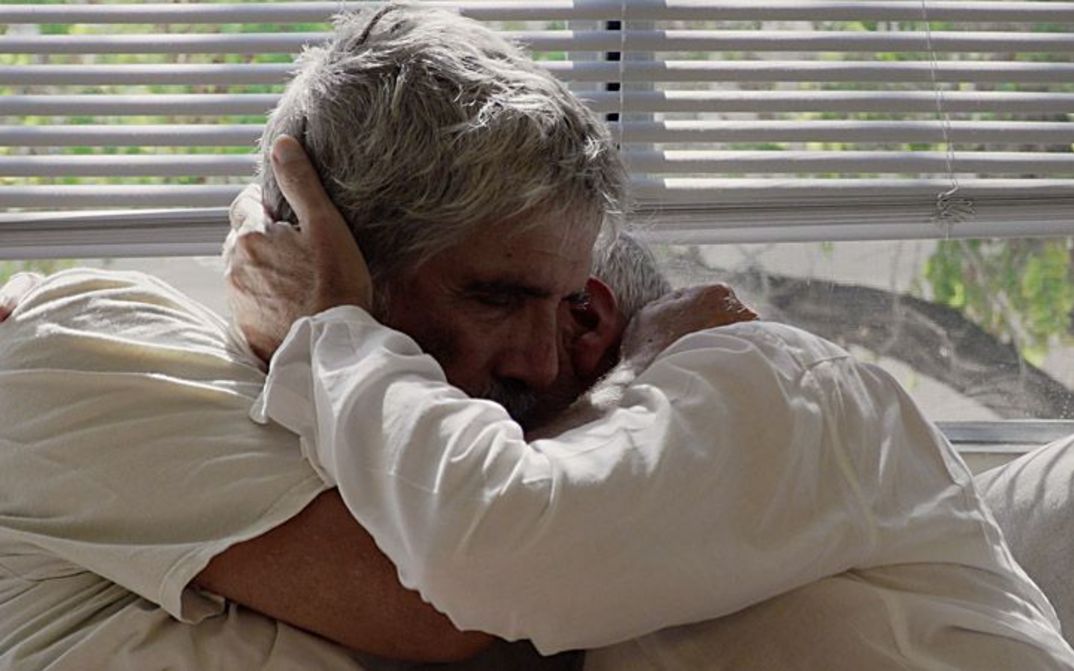 Two elderly men embracing intimately on a couch, both have grey hair and are wearing white shirt. In the background you can see a tree through the window.