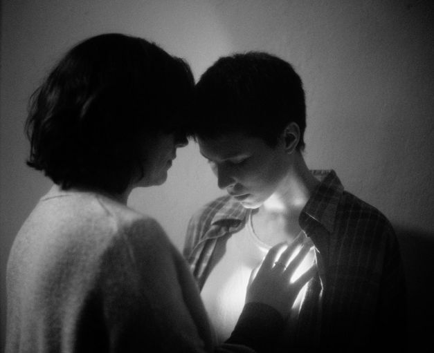 Film still from BATIM by Veronica Nicole Tetelbaum. The black-and-white picture shows two people leaning their heads against each other. The person on the left is holding their hand to the heart of the person opposite.