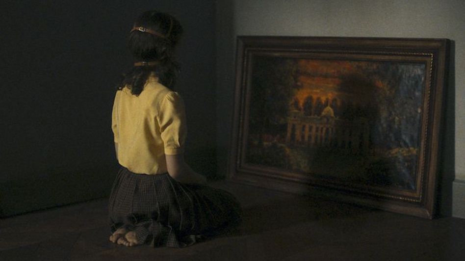 Film still from EARWIG: A girl kneels in front of a painting depicting a castle. She can only be seen from behind, and a kind of mask is strapped around her head with buckles.