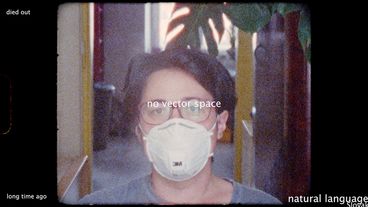 Still aus dem Film „Notes from Eremocene“ von Viera Čákanyová, die Regisseurin selbst sitzt in einem Raum und schaut in die Kamera, sie trägt eine weiße FFP2-Maske.