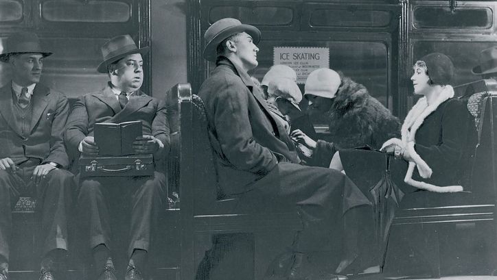Filmstill aus BLACKMAIL: Mehrere elegant gekleidete Menschen sitzen in einem Zug. Auf einem Sitz ist Alfred Hitchcock zu sehen. Er hält ein aufgeschlagenes Buch in den Händen, blickt aber zu den anderen Passagieren.