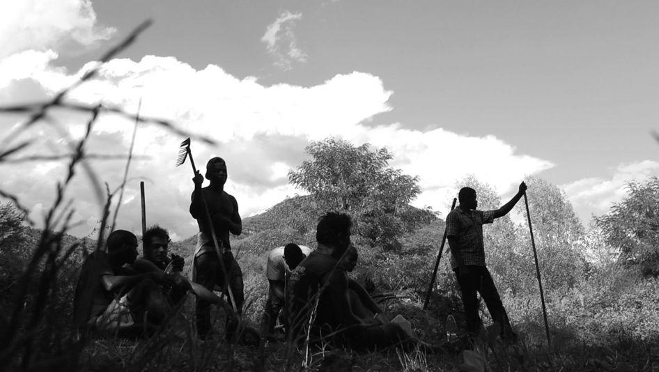 Filmstill aus GEOLOGY OF SEPARATION: Eine Gruppe von Männern steht und sitzt zwischen Bäumen in einer hügeligen Landschaft.