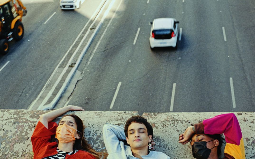 Still from the film "Três tigres tristes" by Gustavo Vinagre. We see the shoulders and heads of three young people laying down on a concrete bridge, while traffic flows beneath them.