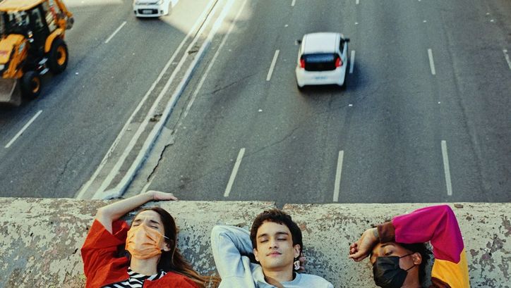 Still from the film "Três tigres tristes" by Gustavo Vinagre. We see the shoulders and heads of three young people laying down on a concrete bridge, while traffic flows beneath them.