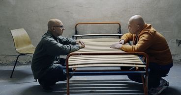 Still from the film "Jaii keh khoda nist" by Tamadon Mehran. Two men are crouching, leaning against an empty bed frame between them, seemingly in conversation with each other.
