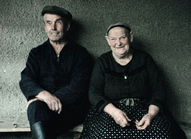 Filmstill aus dem Film "Ein Herbst im Ländchen Bärwalde" von Gautam Bora. Ein Mann mit dunkler Mütze und einer dunklen Jacke sitzt vor einer Wand auf einer Bank neben einer Frau mit einem schwarzen Haarband und einem schwarzen Rock mit weißen Punkten. Beide lächeln.