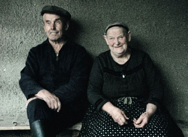 Still from the film "Ein Herbst im Ländchen Bärwalde" by Gautam Bora. A man with a dark cap and a dark jacket is sitting on a bench in front of a wall, next to a women with a black hair ribbon and a black skirt with white dots on it. They are both smiling.