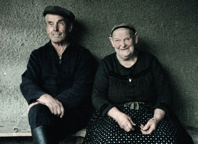 Still from the film "Ein Herbst im Ländchen Bärwalde" by Gautam Bora. A man with a dark cap and a dark jacket is sitting on a bench in front of a wall, next to a women with a black hair ribbon and a black skirt with white dots on it. They are both smiling.