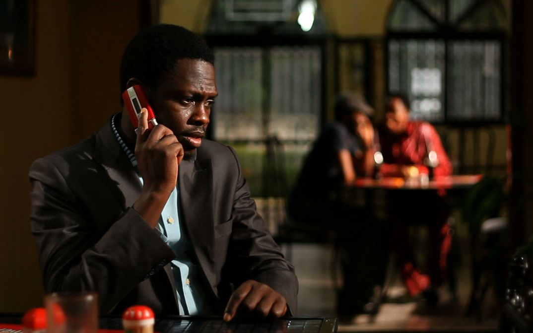 Filmstill aus CONFUSION NA WA: Ein Mann im Anzug am Telefon. Im Hintergrund sieht man unscharf zwei Menschen an einem Tisch sitzen.