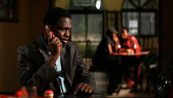 Film still from CONFUSION NA WA: A man in a suit on the phone. In the background you can see two people sitting at a table, out of focus. 