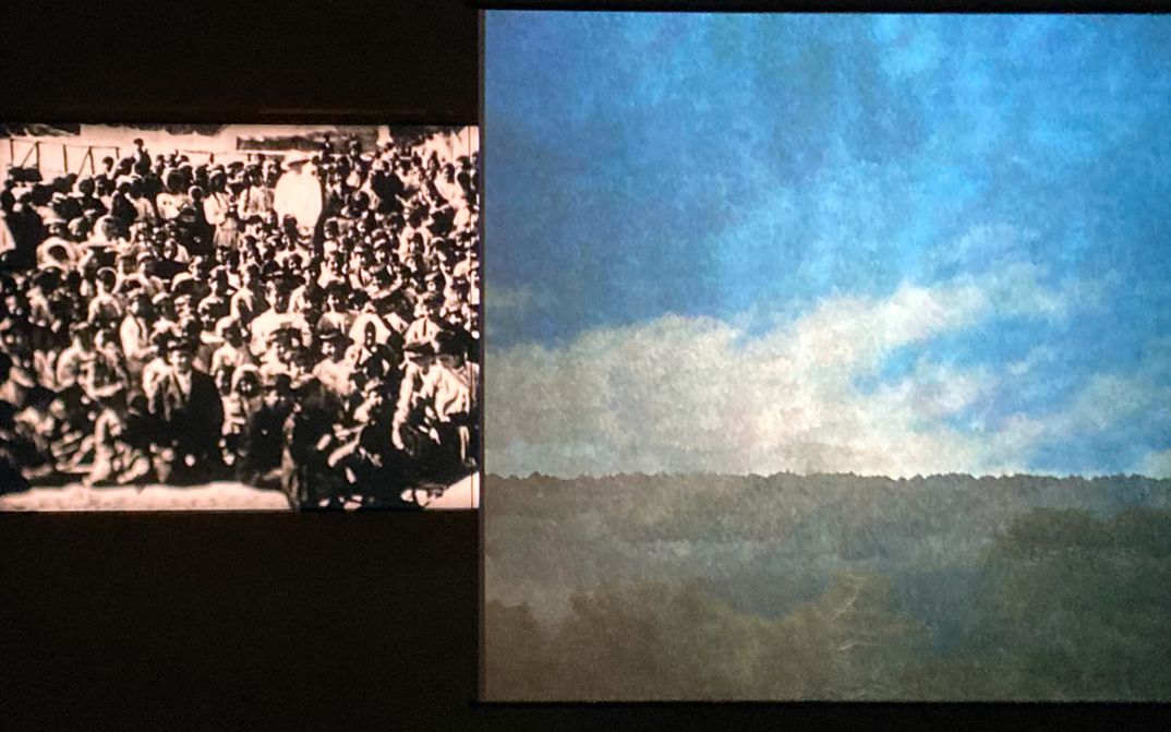Film still from “Industries of Denial, Stage 10: From Musa Dagh to Port Saïd” by Angela Melitopoulos and Kerstin Schroedinger. Two projections hang side by side in the dark room: on the left, a grainy black-and-white archive film showing a densely packed crowd; on the right, a tranquil, colorful landscape image with sky and horizon.