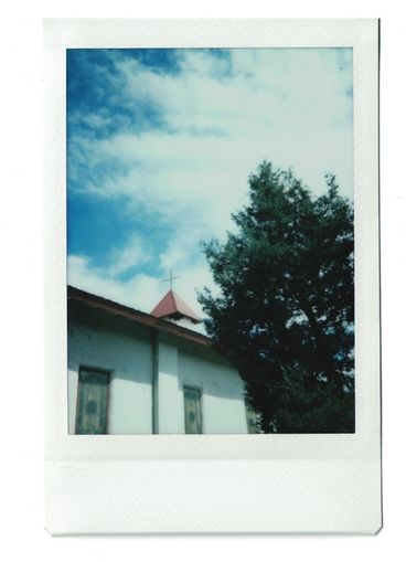 Polaroid of a church seen from below, with a steeple, stained glass windows, and a large green tree.