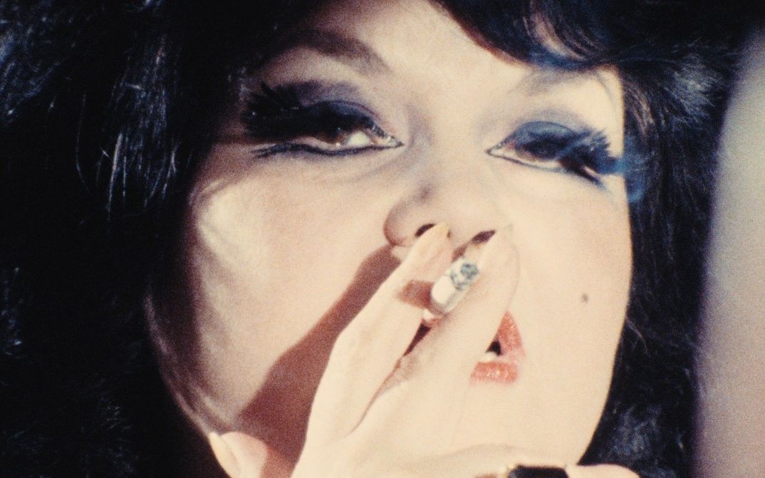 Film still from TALLY BROWN, NEW YORK: Close-up of a dark-haired woman with dark eye make-up smoking a cigarette.