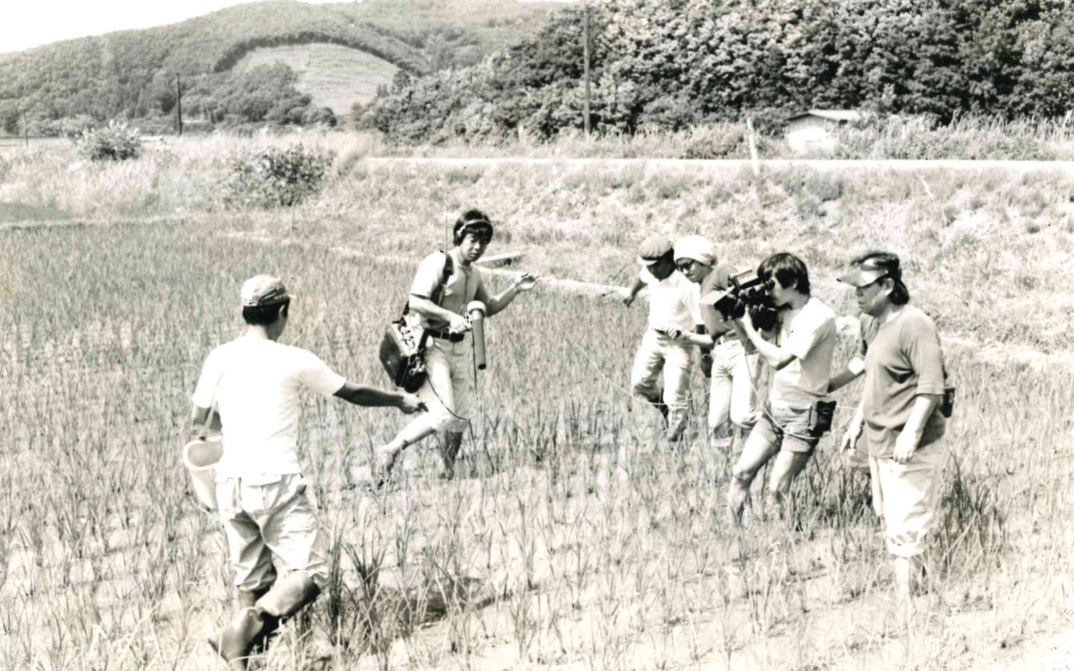 Filmstill aus „Nippon“: Furuyashiki Village: Ein paar Menschen stehen in einem Reisfeld, ein kleines Filmteam nimmt auf.
