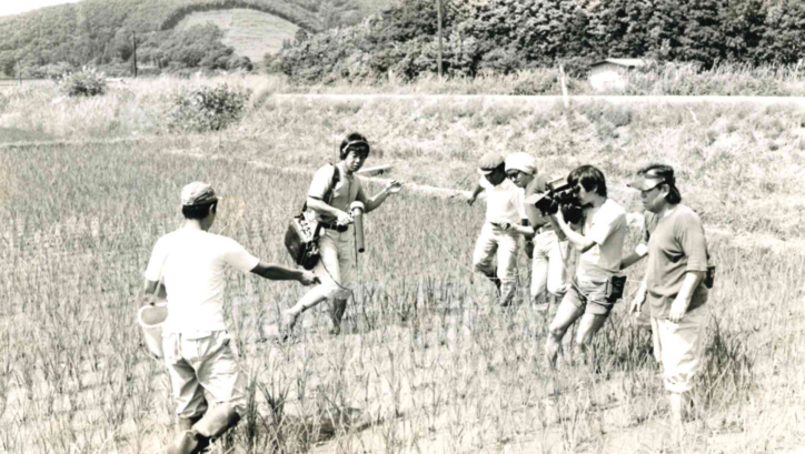 Film still from „Nippon“: Furuyashiki Village: A few people are standing in a rice field, a small film crew is recording.