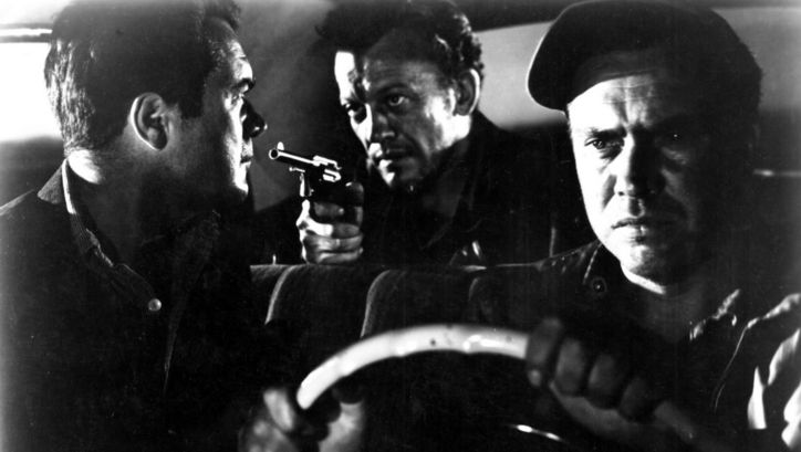 Film still from THE HITCH-HIKER: Three men are sitting in a car. The man in the back seat points a revolver at the man in the passenger seat.