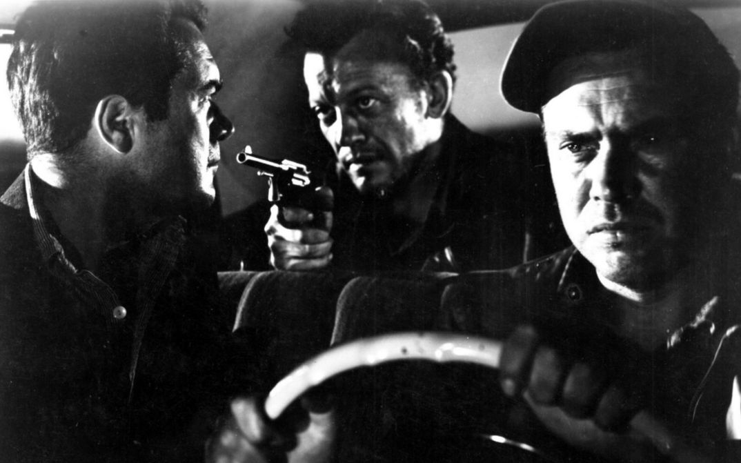 Filmstill aus THE HITCH-HIKER: In einem Auto sitzen drei Männer. Der Mann auf der Rückbank richtet einen Revolver auf den Mann im Beifahrersitz.