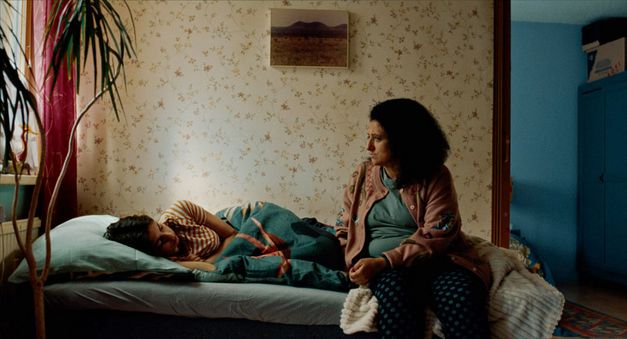 Film still from WENN DU ANGST HAST NIMMST DU DEIN DEIN HERZ IN DEN MUND UND LÄCHELST by Marie-Luise Lehner. It shows a colorful bedroom with a bed on which a young person is lying. A woman is sitting on the edge of the bed. 