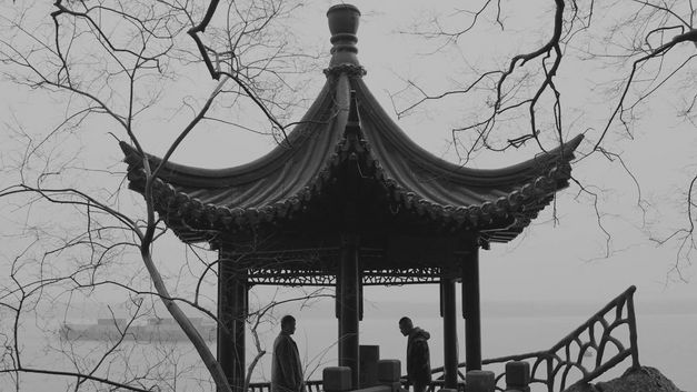 Zwei Personen stehen unter einem chinesischen Pavillon mit geschwungenem Dach am Wasser.