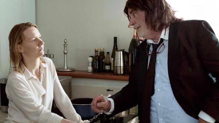Filmstill aus TONI ERDMANN: Ein junge Frau und ein älterer Mann stehen in einer Küche. Sie sind mit Handfesseln aneinander gekettet.