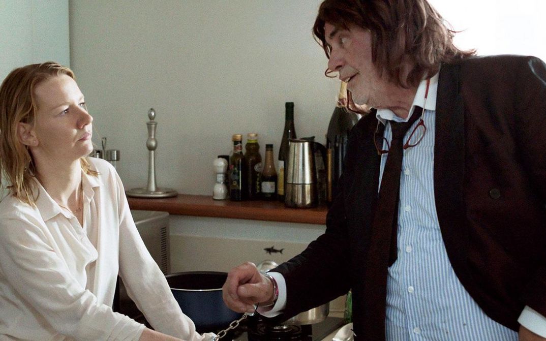 Film still from TONI ERDMANN: A young woman and an older man are standing in a kitchen. They are chained together with handcuffs.