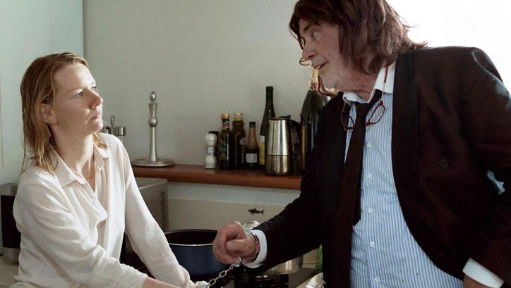 Film still from TONI ERDMANN: A young woman and an older man are standing in a kitchen. They are chained together with handcuffs.