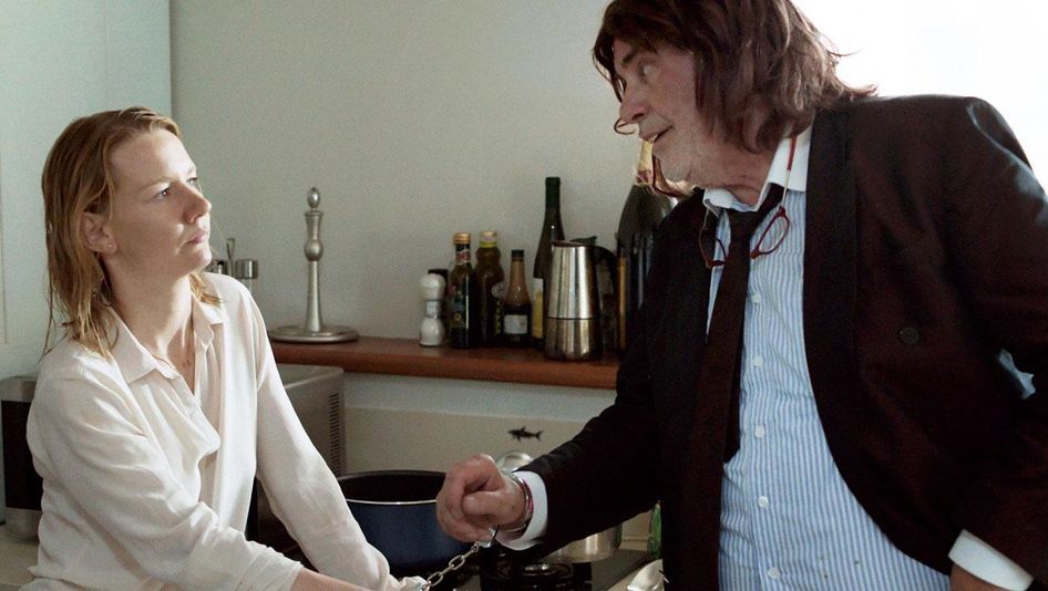Filmstill aus TONI ERDMANN: Ein junge Frau und ein älterer Mann stehen in einer Küche. Sie sind mit Handfesseln aneinander gekettet.