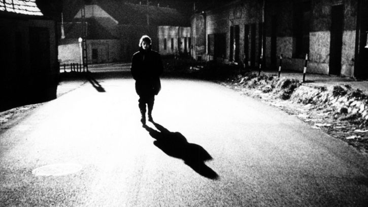 Film still from Werckmeister Harmóniák:  A person is walking alone down a street at night. A streetlight casts a shadow.
