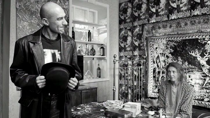 Filmstill aus ETERNAL RETURN/ETERNAL HOMECOMING: Ein Mann und eine Frau sind in einer Wohnung. Sie sitzt am Tisch, die Wand hinter ihr ist voller Poster.