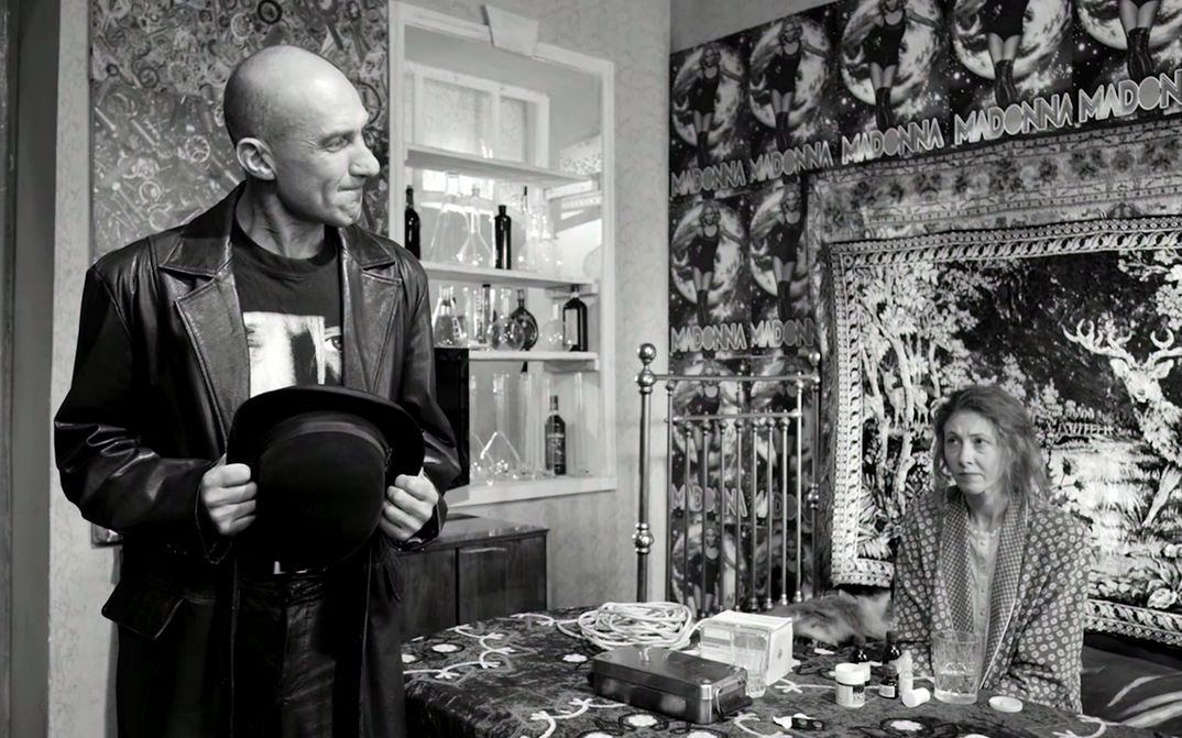 Filmstill aus ETERNAL RETURN/ETERNAL HOMECOMING: Ein Mann und eine Frau sind in einer Wohnung. Sie sitzt am Tisch, die Wand hinter ihr ist voller Poster.