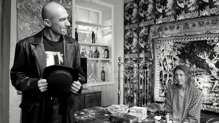 Film still from ETERNAL RETURN/ETERNAL HOMECOMING: A man and a woman are in a flat. She is sitting at the table, the wall behind her is full of posters.