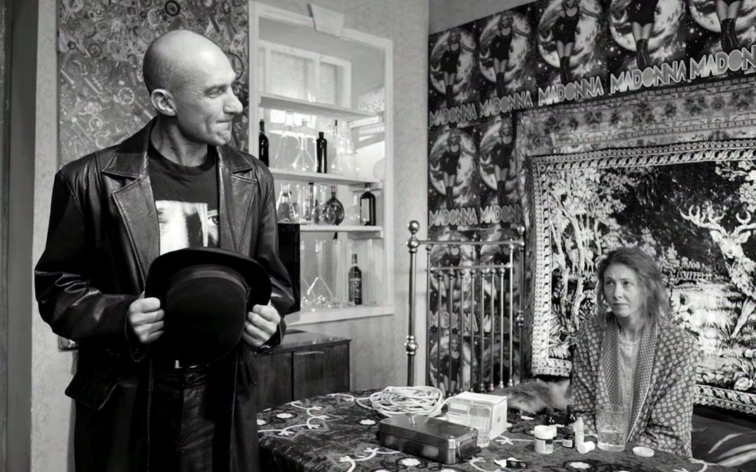 Film still from ETERNAL RETURN/ETERNAL HOMECOMING: A man and a woman are in a flat. She is sitting at the table, the wall behind her is full of posters.