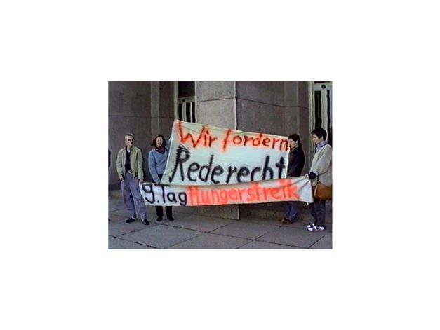 Film still from Anna Zett’s film “Es gibt keine Angst (Afraid Doesn’t Exist)”. People holding a banner with the inscription "We demand the right to speak. 9th day of hunger strike" in front of the entrance of a building. (Banner in german, author