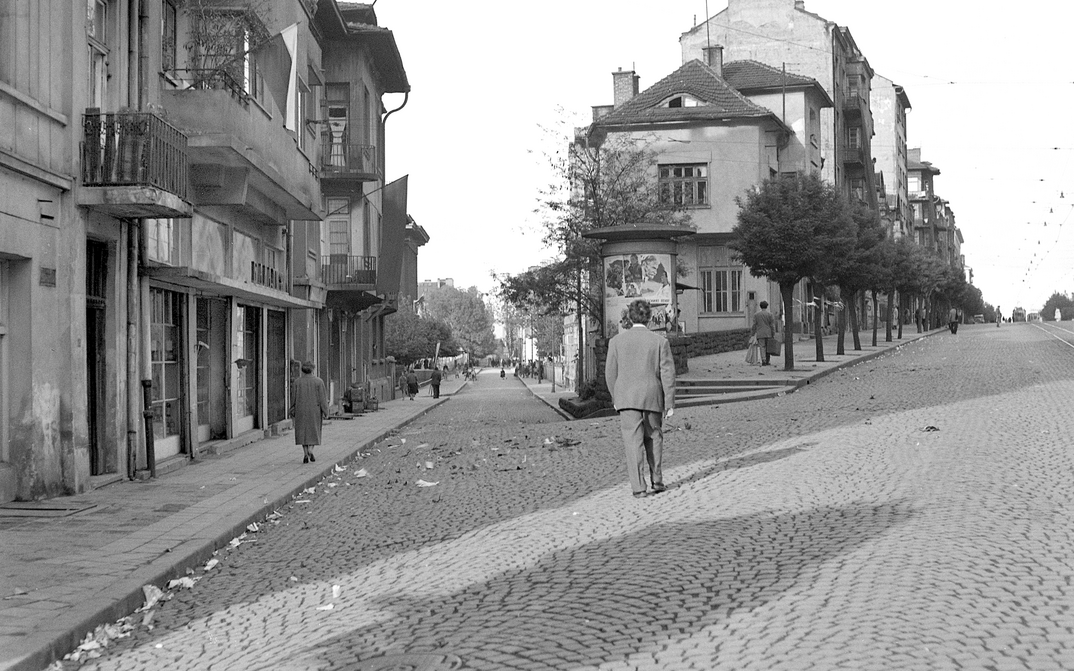 Film still from LIFE FLOWS SLOWLY BY: You see a street scene with several people, all with their backs turned to us.