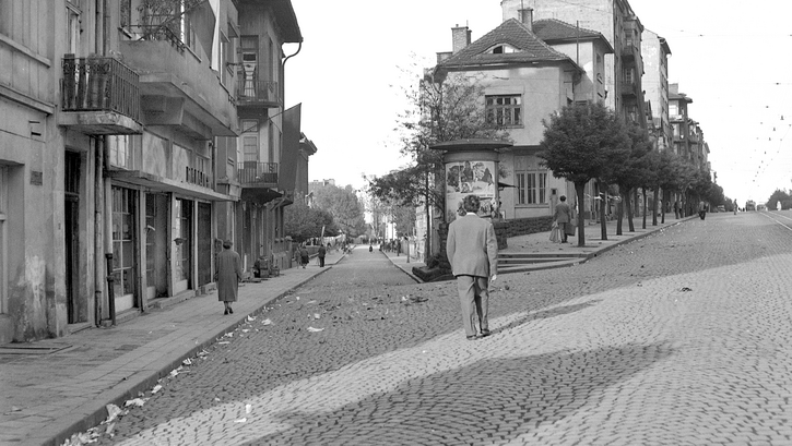 Film still from LIFE FLOWS SLOWLY BY: You see a street scene with several people, all with their backs turned to us.